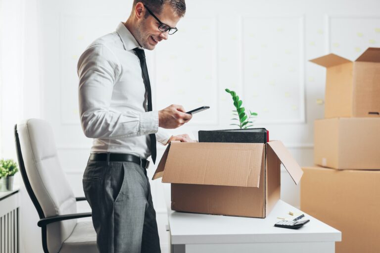 Homme d’affaires vérifiant son smartphone près d’un carton en cours de déménagement, bureau et cartons empilés en arrière-plan. checklist déménagement prêt à télécharger.