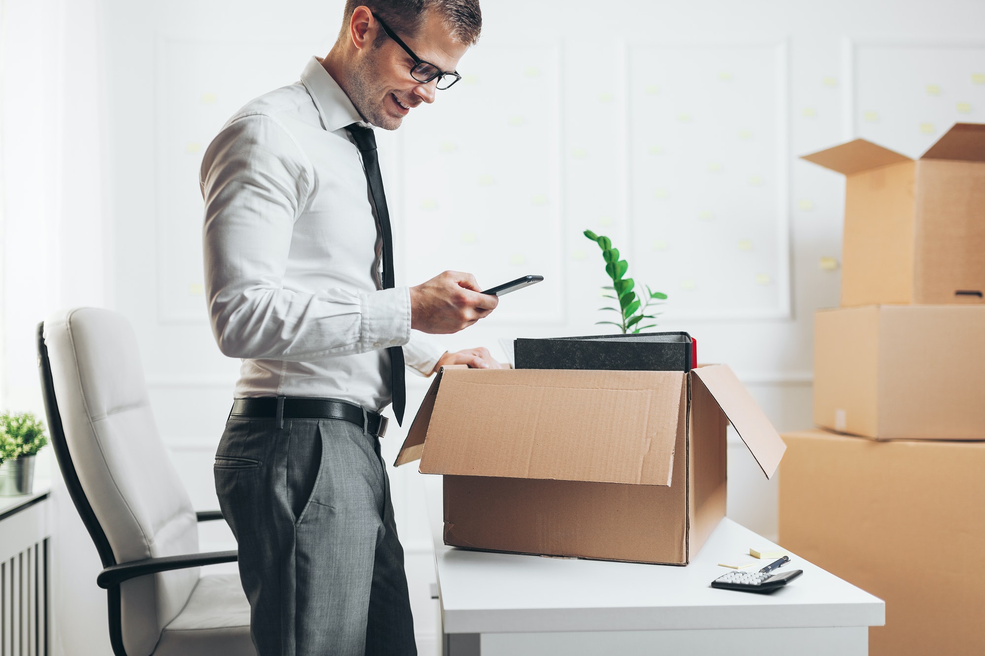 Homme d’affaires vérifiant son smartphone près d’un carton en cours de déménagement, bureau et cartons empilés en arrière-plan. checklist déménagement prêt à télécharger.